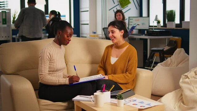 Pleased multiracial business partners sitting on couch in modern office shaking hands after signing partnership contract. Diverse team being happy for successful negotiations in start up company.