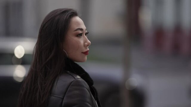 Portrait of a beautiful and spectacular Asian woman with red lipstick on her lips in a stylish image, walking with a confident gait along a city street against the background of cars.