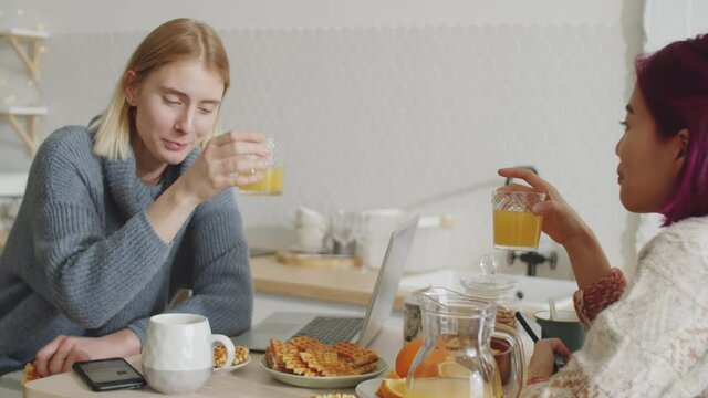 Young Caucasian and Asian female flatmates sitting at kitchen table, drinking orange juice, eating waffles and chatting in the morning at home