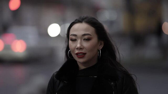 Portrait of a beautiful and spectacular Asian woman with red lipstick on her lips in a stylish image, walking with a confident gait along a city street against the background of evening lights.