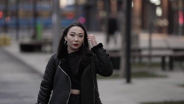 The view of a beautiful and spectacular Asian woman with red lipstick on her lips in a stylish image, walking with a confident gait along a daytime city street in the background.