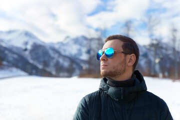 Fototapeta na wymiar Stylish young man in down jacket and sunglasses standing in front of ski lifts and snow mountains panorama covered in snow. Confidently looking away on territory of winter resort near Sochi, Russia 