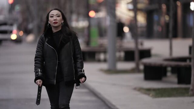 Front view of a beautiful and spectacular Asian woman with red lipstick on her lips in a stylish image, walking with a confident gait along a daytime city street in the background.