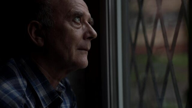 Portrait of an elderly bald man in a shirt carefully looking out the window.