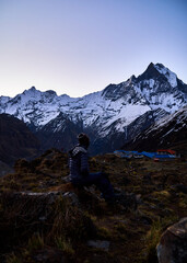 Fototapeta premium Person sitting by a tent at dusk gazing at the snow-capped Annapurna and Machapuchare peaks in the Nepal Himalayas