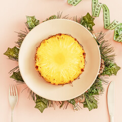 The concept of a diet after the Christmas holidays. Food for weight loss, taking care of the figure. Pineapple on a plate decorated with Christmas decorations with measuring tape on a pink background