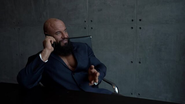 a muscular, bald, bearded man in a black shirt sits at a Desk and uses a mobile phone, talking on it and smiling.