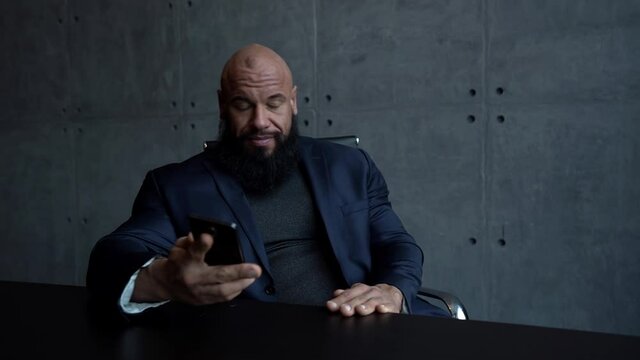 muscular Jock a bald man sits at home at a table and uses a mobile phone, talking on it and smiling.