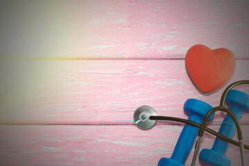 A heart with a stethoscope lies on a wooden background next to dumbbells. Healthy heart
