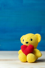 hand make yarn red heart put on yellow teddy bear in front of white pot and green ornamental plants on wooden table and blue background. Concept of valentines day.
