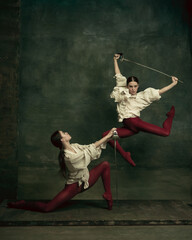 Fight. Two young female ballet dancers like duelists with swords on dark green background. Caucasian models dancing together. Ballet and contemporary choreography concept. Creative art photo.