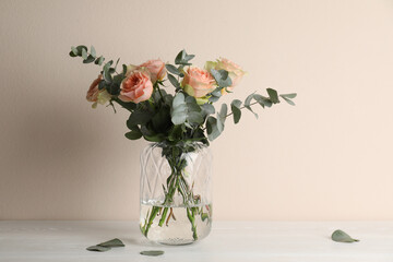 Vase with bouquet of beautiful roses on white wooden table near beige wall