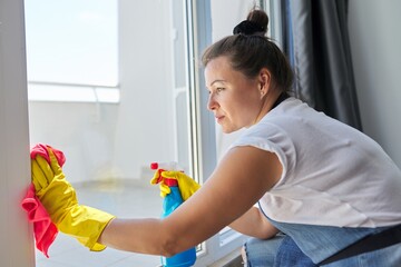 House cleaning, mature woman in rubber gloves wearing an apron washing windows