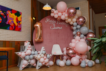 Beautiful festive decorations, pink and grey balloons arch, wooden stars, white chair and number one balloon on wooden round background. Little 1 year old girl birthday party photo zone.
