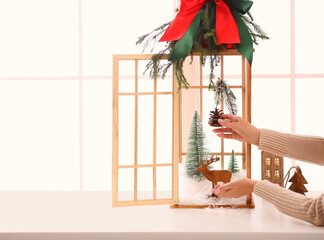 Woman creating Christmas composition inside of vintage wooden lantern on window sill indoors, closeup. Space for text