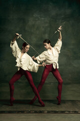 Fototapeta na wymiar Contrast. Two young female ballet dancers like duelists with swords on dark green background. Caucasian models dancing together. Ballet and contemporary choreography concept. Creative art photo.