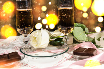 Against the backdrop of glittering lights, there are two glasses of champagne, chocolate and a white rose.