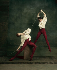 Bright. Two young female ballet dancers like duelists with swords on dark green background. Caucasian models dancing together. Ballet and contemporary choreography concept. Creative art photo.