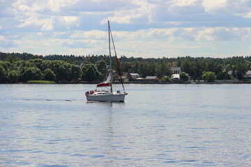 One white sailing yacht with mast without sail floats on Moscow Region water reservoir with green forest trees on far shore at summer day on blue sky with clouds background, river tourism recreation