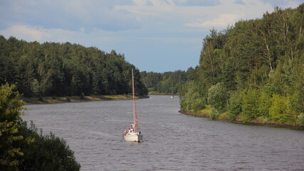 One white sailing yacht with mast  without sail floating on Moscow Canal river turn with green forest trees on shores at summer day on blue sky background, water tourism recreation