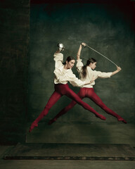 Fight. Two young female ballet dancers like duelists with swords on dark green background. Caucasian models dancing together. Ballet and contemporary choreography concept. Creative art photo.