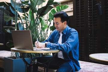Ethnic distance worker writing on paper near laptop in cafeteria