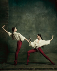 Attacking. Two young female ballet dancers like duelists with swords on dark green background. Caucasian models dancing together. Ballet and contemporary choreography concept. Creative art photo.