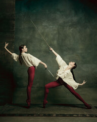 Attacking. Two young female ballet dancers like duelists with swords on dark green background. Caucasian models dancing together. Ballet and contemporary choreography concept. Creative art photo.