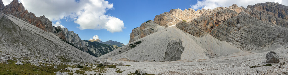 Fototapeta na wymiar A panoramic view on a stony valley in Italian Dolomites. There are high and sharp mountains around. In the back the mountains are overgrown with green plants. Remote and raw landscape. Sunny day