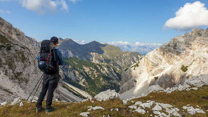 Fototapeta premium A man with a big hiking backpack admiring the view on a stony valley in Italian Dolomites. There are high and sharp mountains around. Remote and raw landscape. Sunny day. Freedom and serenity