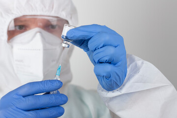 Male nurse with protective coverall clothing, with face mask and blue gloves, holding syringe with needle and covid-19 vaccine vial.