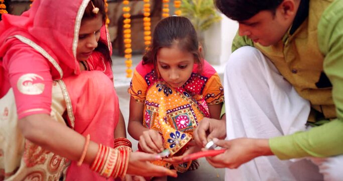 Couple with daughter wearing colorful traditional dress decorating floor in Rangoli- pattern with flower petals, brass bowl with floating candles oil lamps for Diwali- Hindu festival of lights