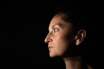 Dark profile portrait of a very dimly lit woman. The woman is looking straight ahead and transmits...