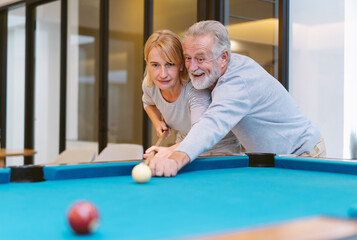 Cheerful senior couple have fun in living room and husband teaching his wife for playing snooker...