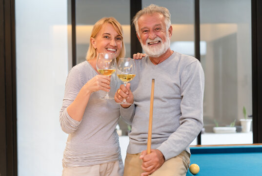 Happy Senior Couple Having Fun In Living Room Playing Billiards Together And Drinking Wine And Looking At Camera. Happy Retirement Concept.