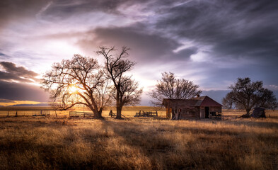 An old farmhouse on the eastern plains of Colorado in a rural setting at sunset. The sky is dramatic with wispy clouds. The old house if falling apart and abandoned.  - obrazy, fototapety, plakaty
