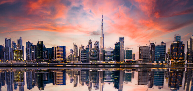 Stunning panoramic view of the Dubai skyline at sunset with buildings and skyscrapers reflected on a silky smooth water flowing in the foreground. Dubai, United Arab Emirates.
