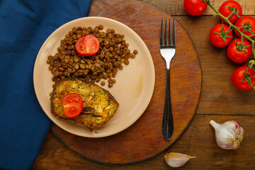 A plate of fish with lentils and tomatoes on a round stand with a fork on a blue napkin next to cherry tomatoes and garlic.