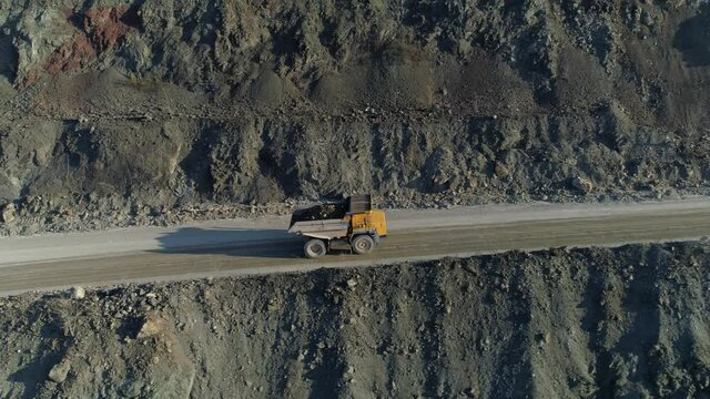Drone flight together with yellow modern truck transporting mining ore. Heavy loaded vehicles are driving along dusty serpentine road up. Mining industry, abstract industrial landscape. Cement quarry