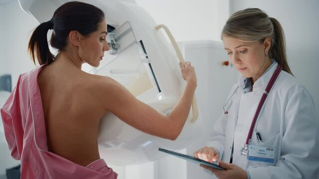 Friendly Female Doctor with Tablet Computer Explains Mammogram Procedure to a Topless Adult Female Patient Undergoing Mammography Scan. Healthy Woman Does Cancer Prevention Routine in Hospital Room.