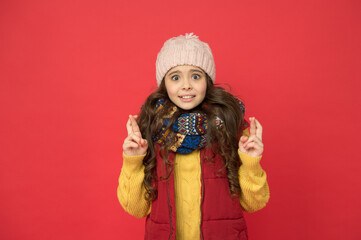 surprised pretty kid girl in puffer waistcoat with knitted hat and sweater hold crossed fingers,...