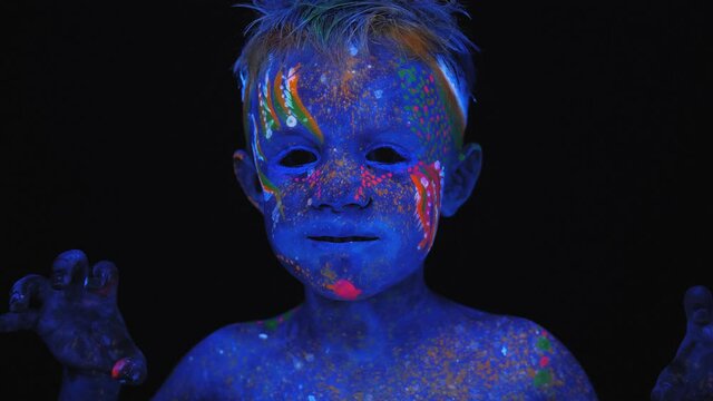 A close-up of a small boy's face with ultraviolet drawings on his body and face, he raises his hands and growls.