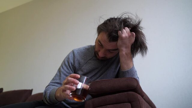 Young man suffering from strong headache or migraine sitting on sofa with whisky glass, millennial guy feeling intoxication and pain touching aching head, morning after hangover concept