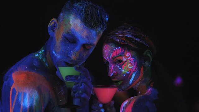 A couple with UV body art dancing with glasses of colorful glowing cocktails in the dark under the light of fluorescent lamps. Art design of couple disco dancers posing in UV.