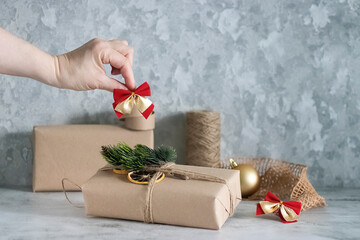 Christmas and New Year concept. Gift in kraft paper with a rope, pieces of decorative paper, Christmas decorative ornaments and a female hand with a red decorative bow. Soft focus.
