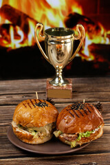 Two freshly fried burgers lie on a plate and a wooden table against the background of a fireplace. Juicy fried dinner in front of fireplace.
Two burgers on a plate with a golden cup