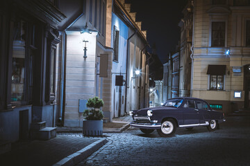 A vintage 1960s Volga car making a right turn on an old cobblestone street between classicist houses in the night - obrazy, fototapety, plakaty