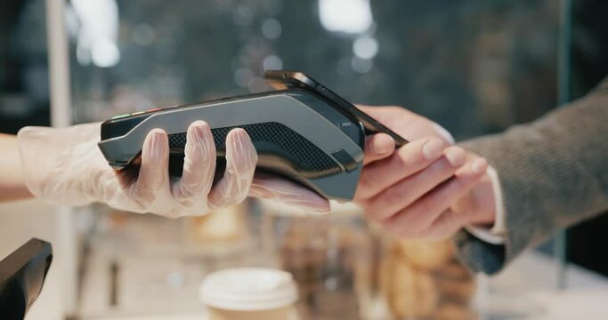 Close-up of female barista worker holding card machine in gloves. Male customer using NFC contactless payment technology. Coffee house. Cafeteria services.