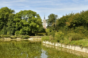 Fototapeta na wymiar L'Etang des Tanneurs entouré de végétation luxuriante laissant entrevoir le clocher de l'église St-Nicolas au parc d'Enghien en Hainaut 