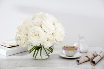 A bouquet of white roses in a glass vase on a table with two cups of coffee, a kettle and a milk jug.